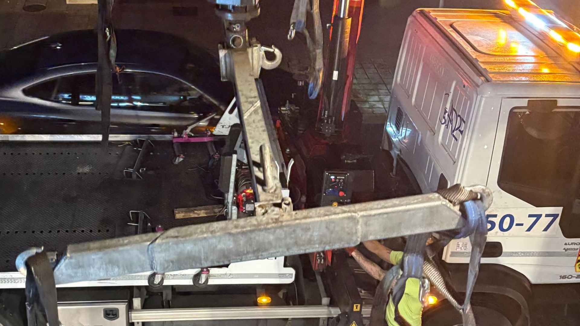 Une dépanneuse charge une voiture sombre sur son plateau la nuit ; l'opérateur de la dépanneuse porte un gilet de sécurité.