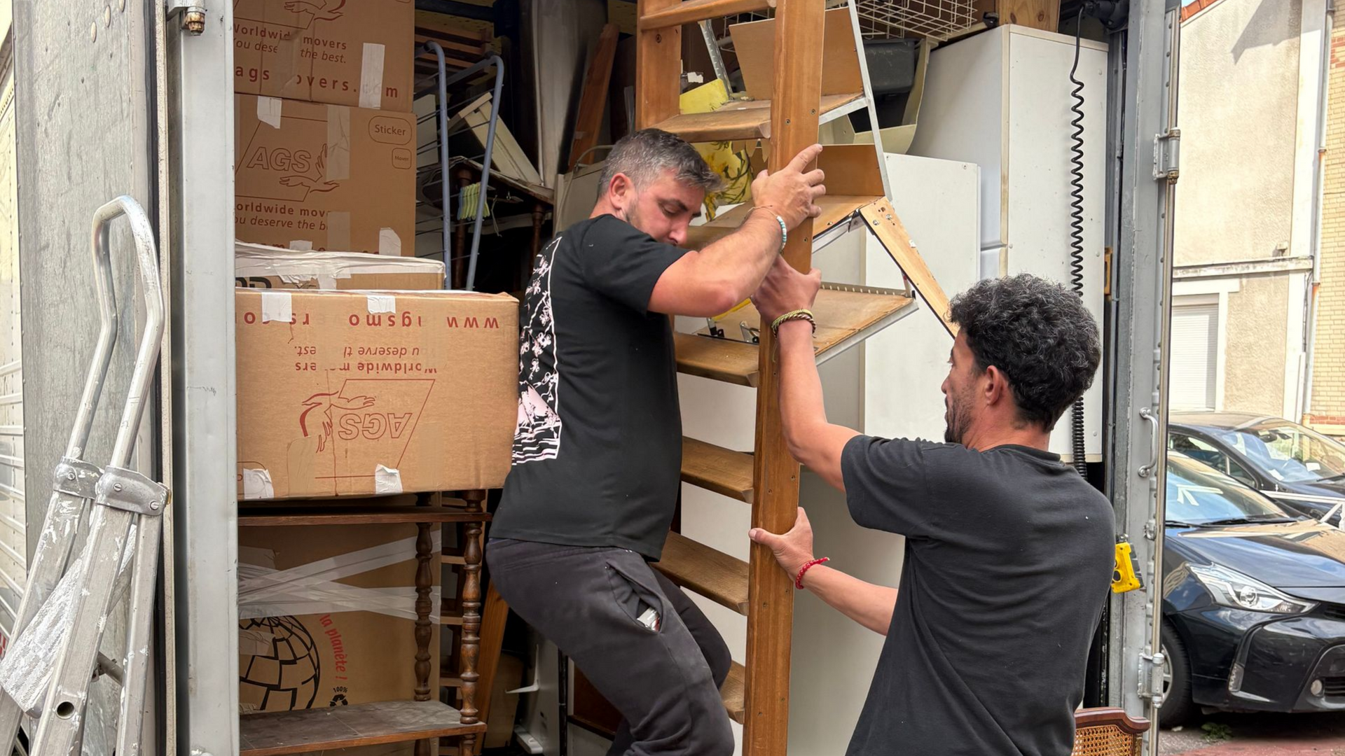 Deux personnes chargent une étagère en bois dans un camion de déménagement, des cartons et des meubles sont visibles.