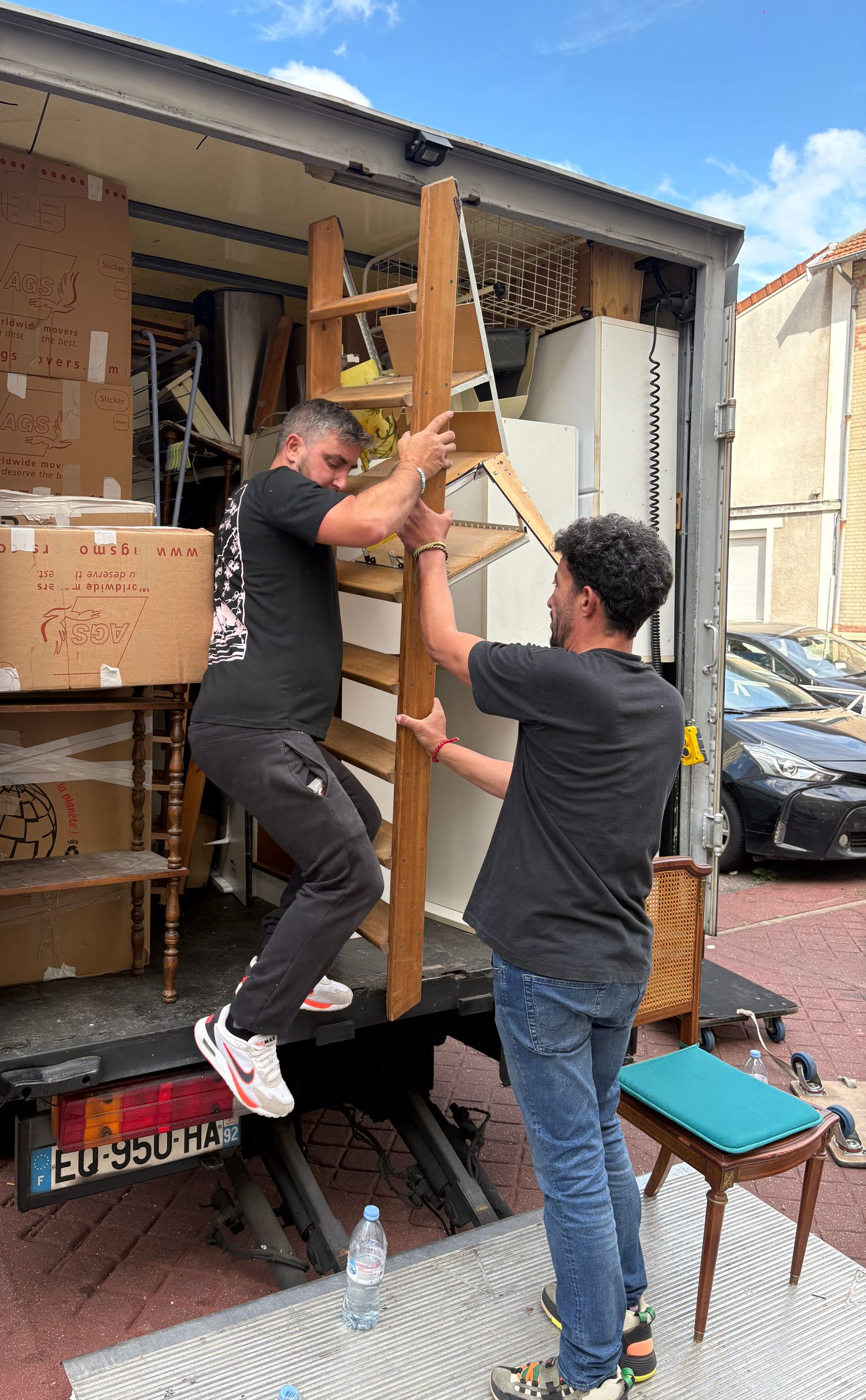 Deux hommes chargent des meubles dans un camion de déménagement ; l'un monte à bord, l'autre stabilise une échelle.