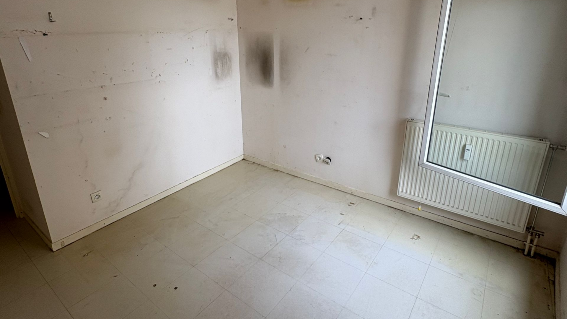 Pièce vide avec des murs blanc cassé, un sol pâle et un radiateur.