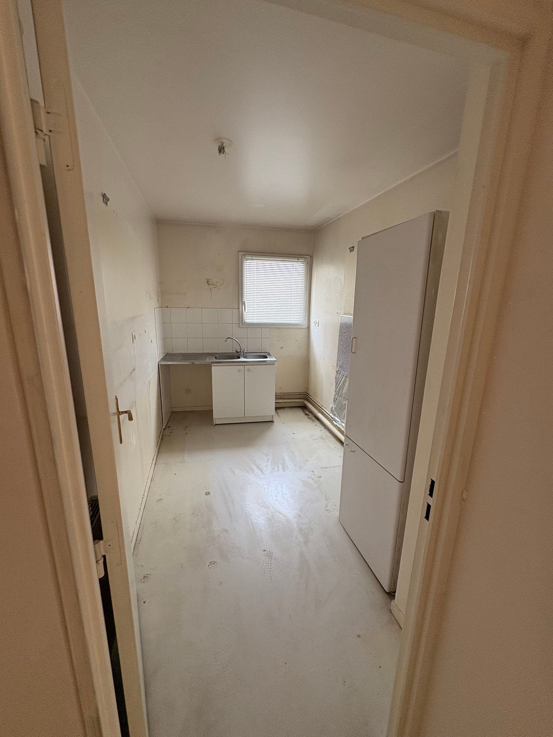 Petite cuisine vide vue depuis la porte ; murs, comptoir et armoires blancs ; stores sur la fenêtre.