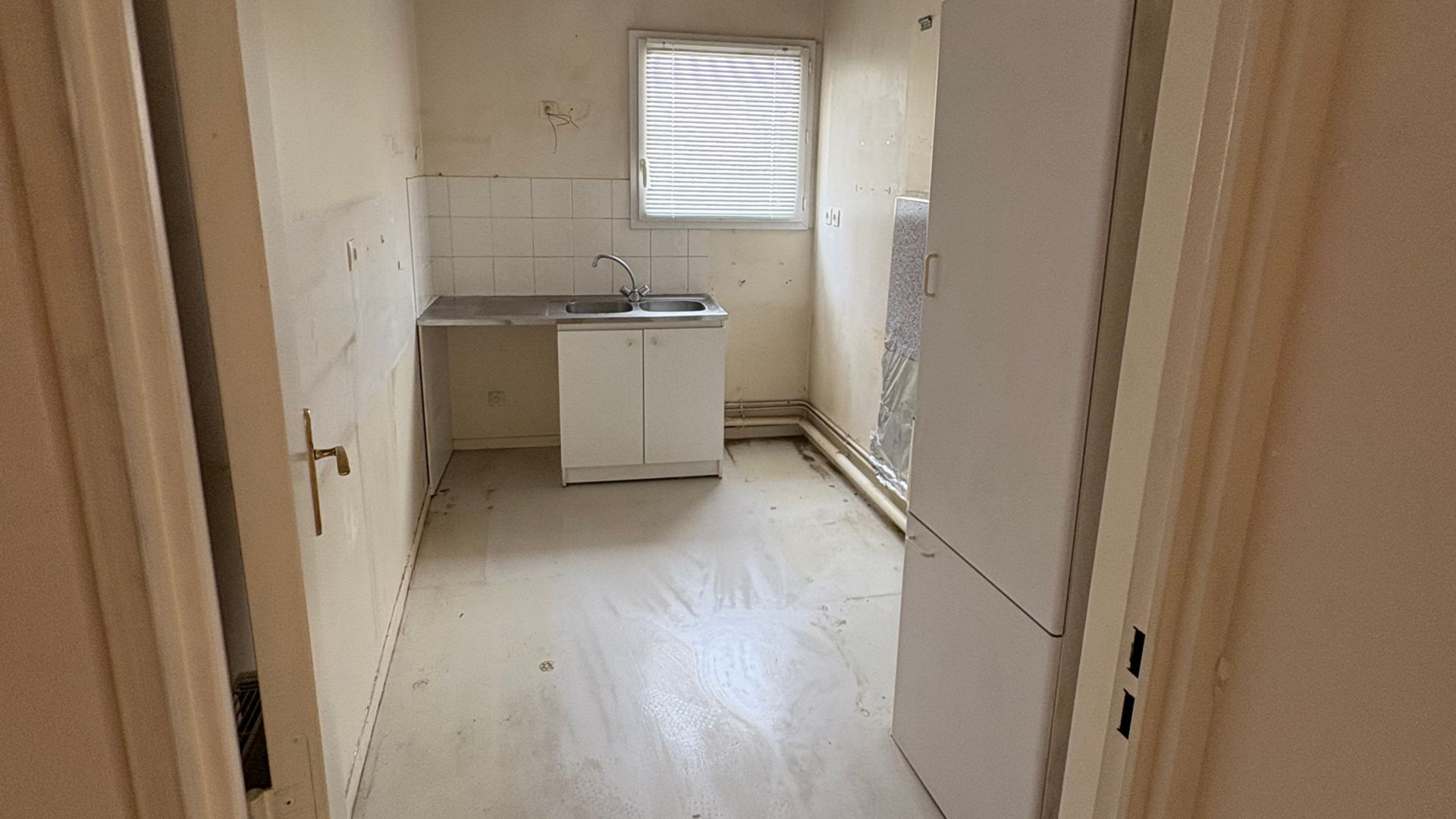 Petite cuisine vide avec un évier, un placard et une fenêtre ; papier peint qui se décolle sur les murs.