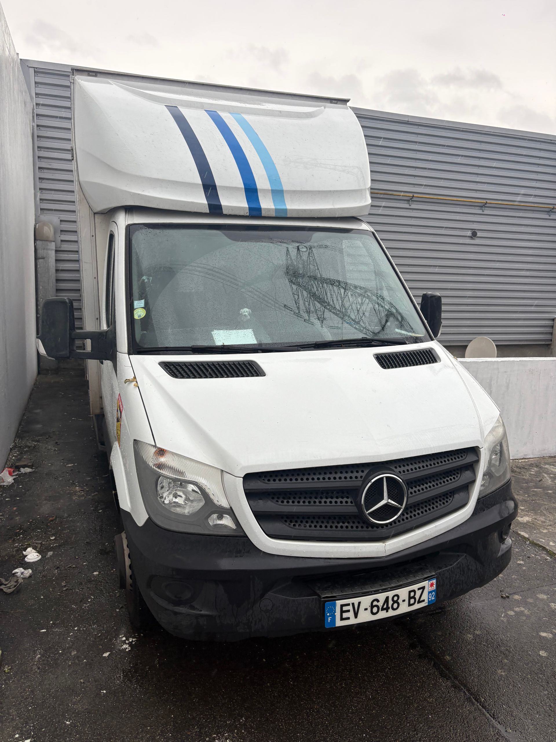Camionnette de livraison Mercedes blanche à rayures bleues, garée à l'extérieur.