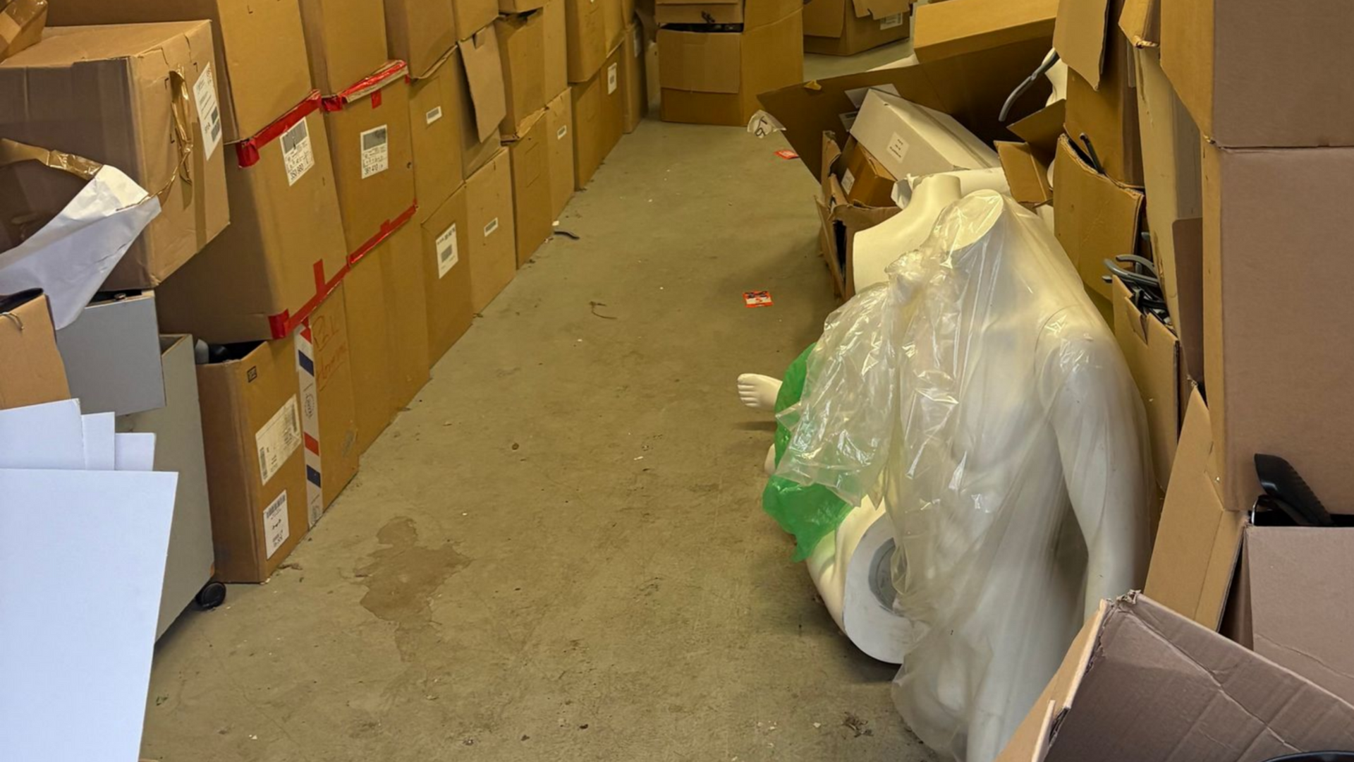 Pièce de rangement étroite avec des cartons tapissant les murs, un mannequin dans un sac au centre.