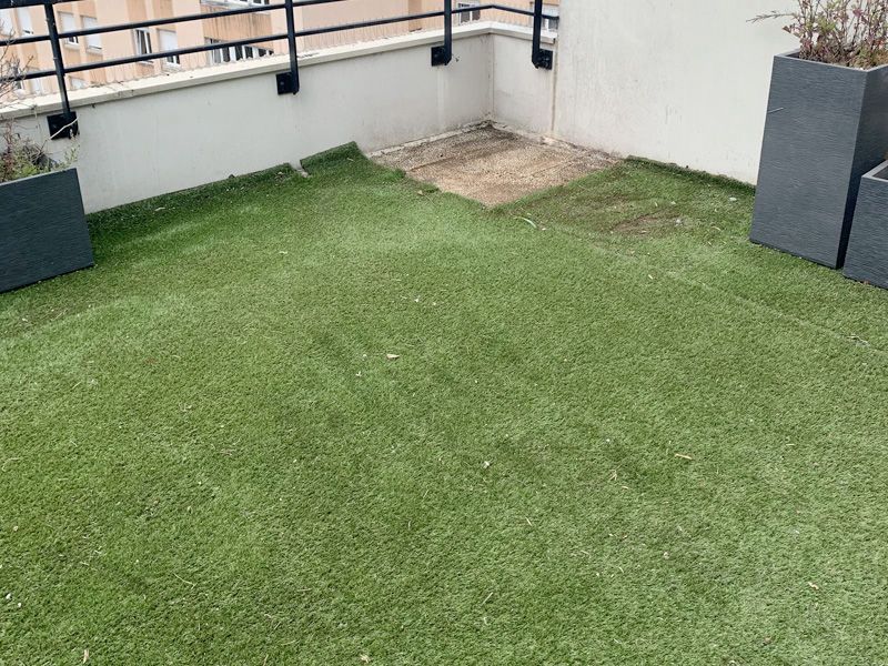 Balcon avec gazon artificiel vert, jardinières et mur blanc, sous une balustrade métallique.