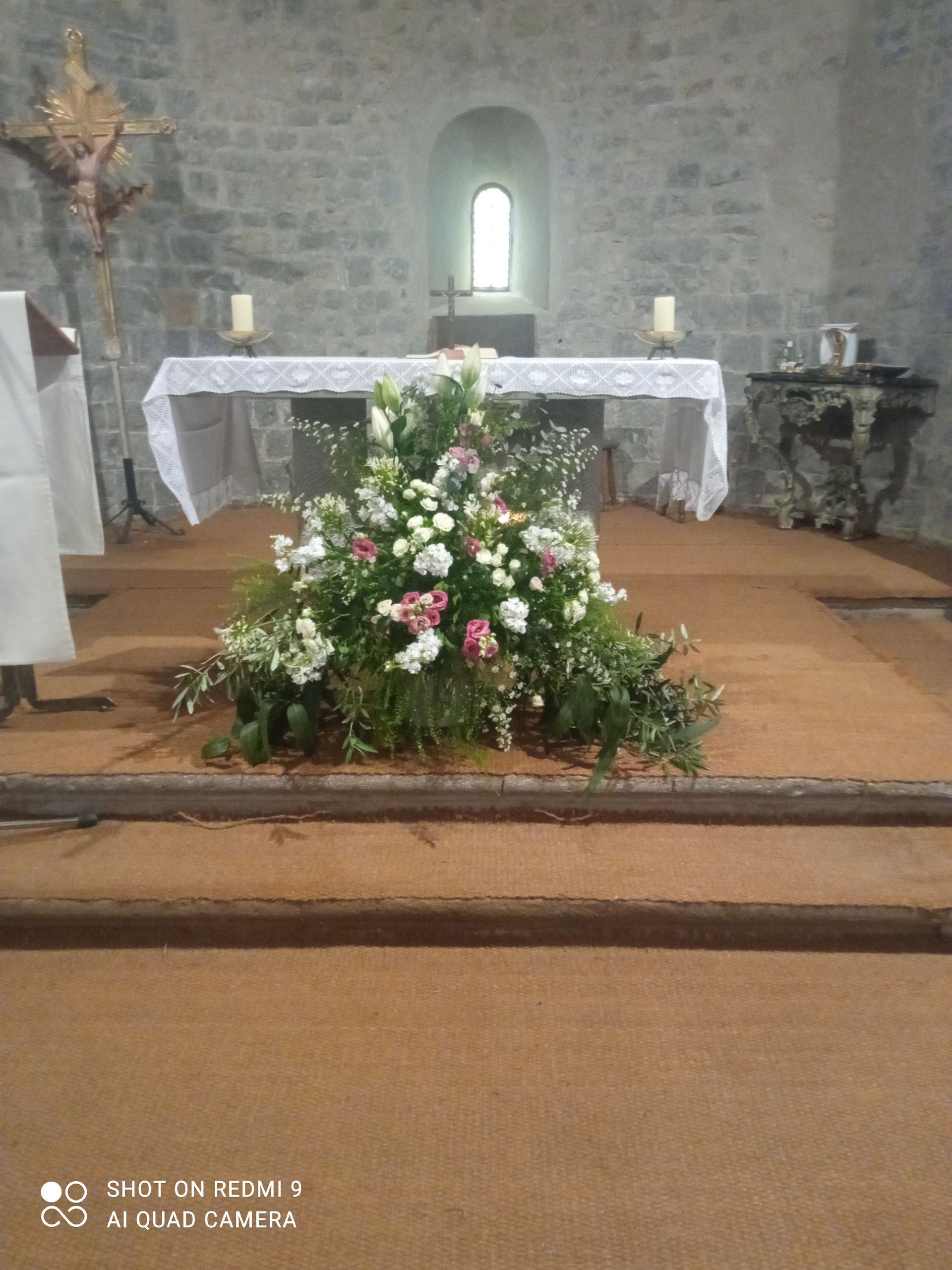 Composition florale sur l'autel d'une église en pierre. Fleurs blanches, roses et vertes, avec une petite fenêtre.