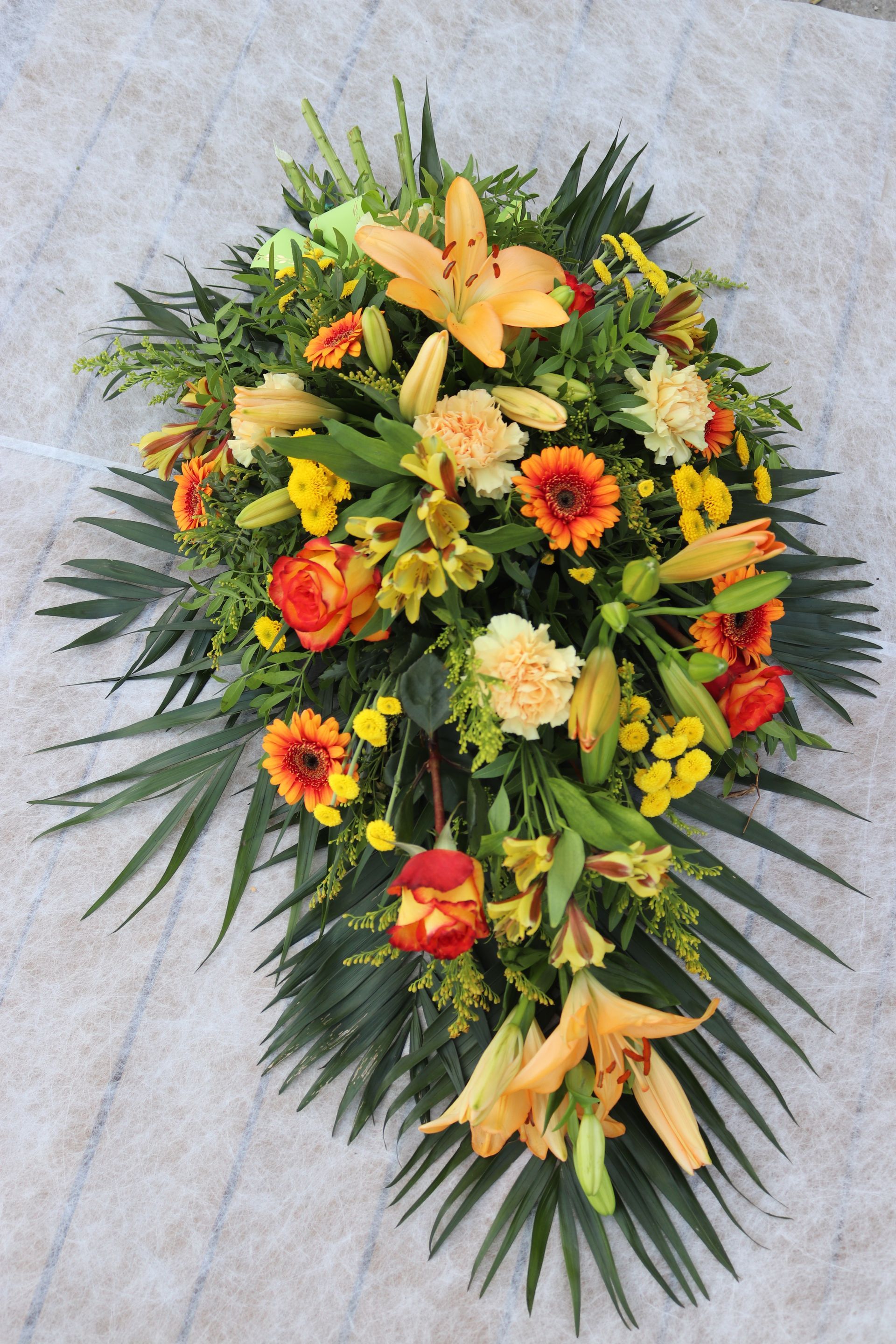 Composition florale avec des lys orange, des gerberas et de la verdure sur un fond clair.