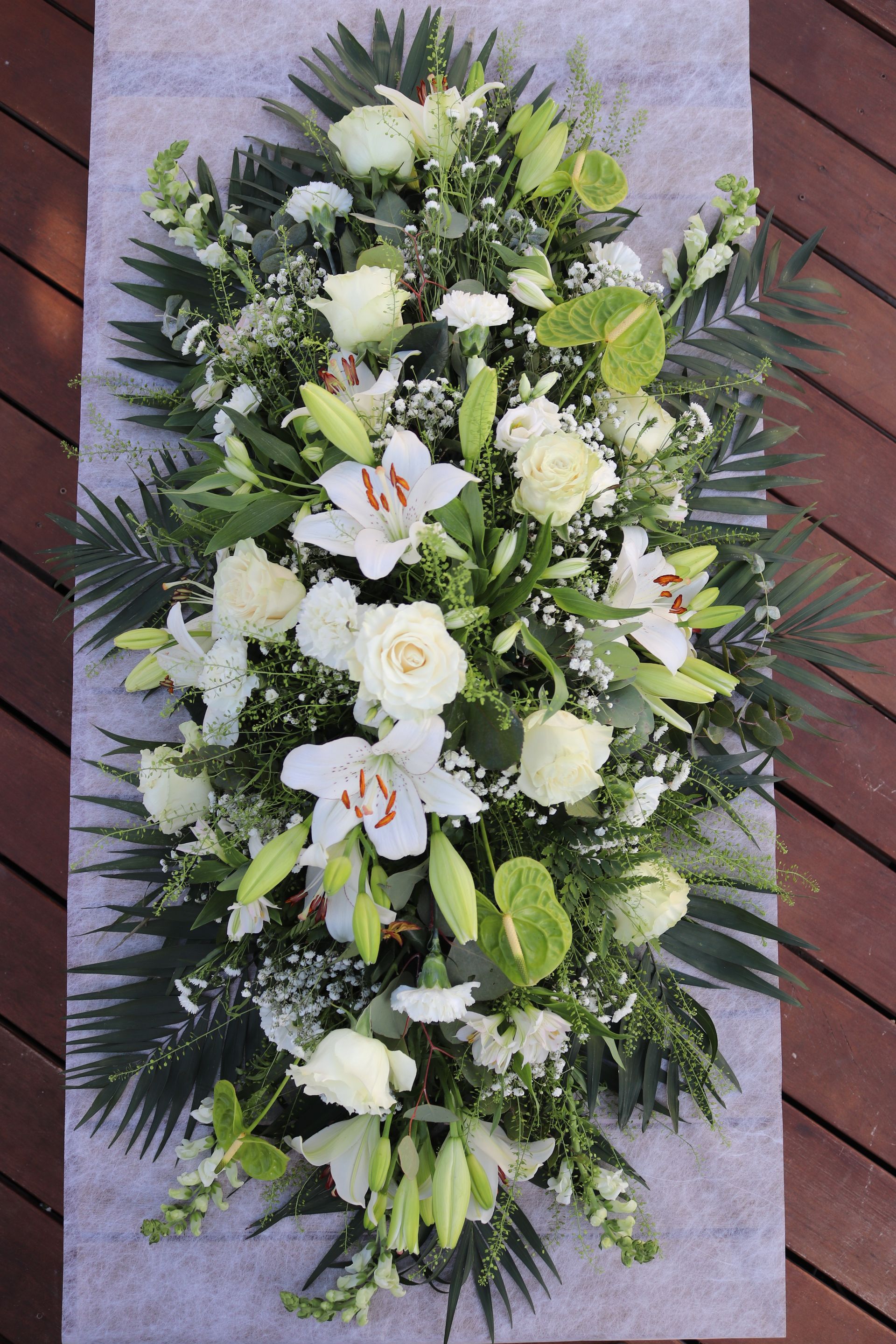 Composition florale blanche pour cercueil sur fond gris clair, avec des lys, des roses et de la verdure.