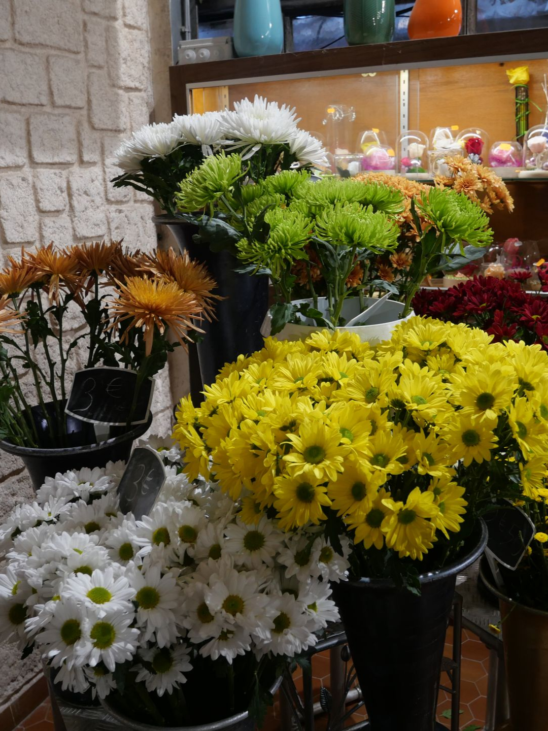 Des chrysanthèmes de couleurs variées sont présentés dans une boutique de fleurs. Des fleurs jaunes, blanches, vertes, oranges et rouges sont disposées dans des vases noirs.