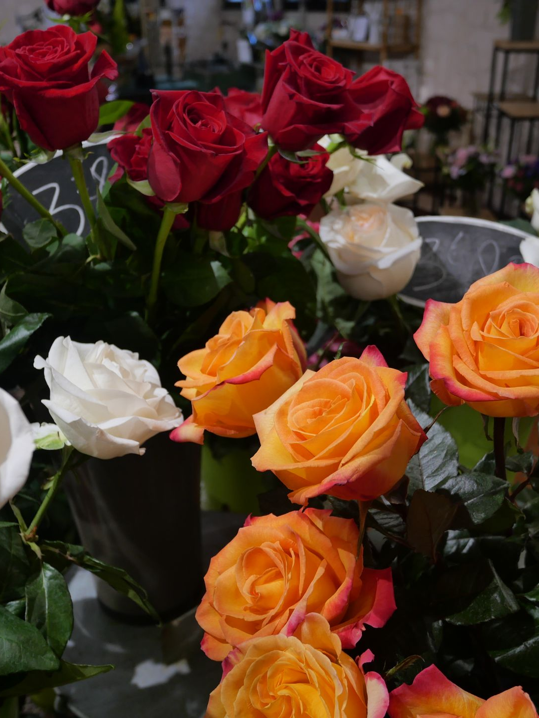 Gros plan sur des bouquets : roses rouges, oranges et blanches avec feuillage vert. Ambiance boutique.