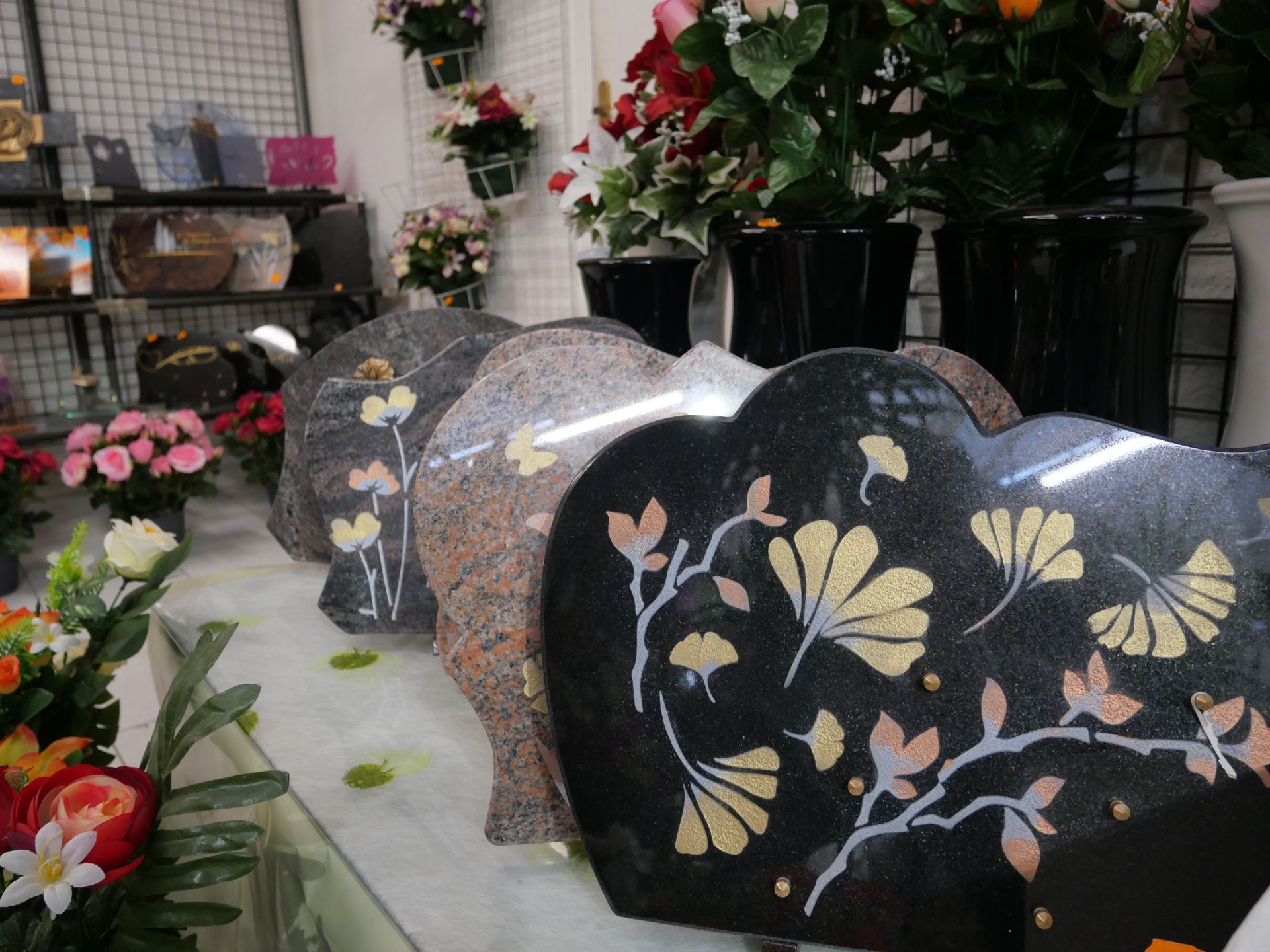 Des pierres tombales en granit en forme de cœur, ornées de motifs floraux, sont exposées dans un magasin, entourées de fleurs.