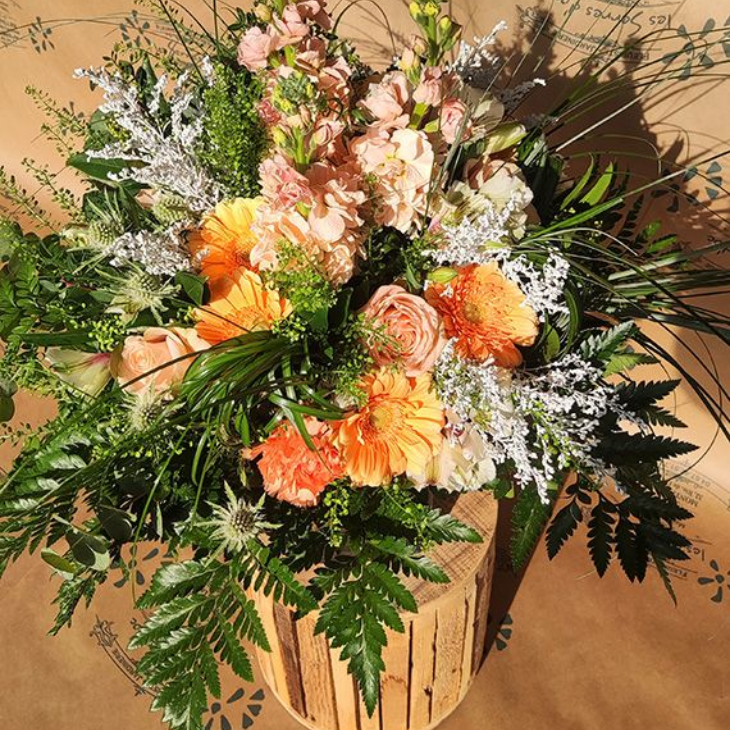 Bouquets de fleurs pêche, orange et blanc avec de la verdure, disposés sur une souche de bois.