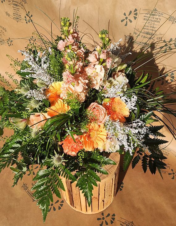 Bouquets de fleurs pêche, orange et blanc avec de la verdure, disposés sur une souche de bois.