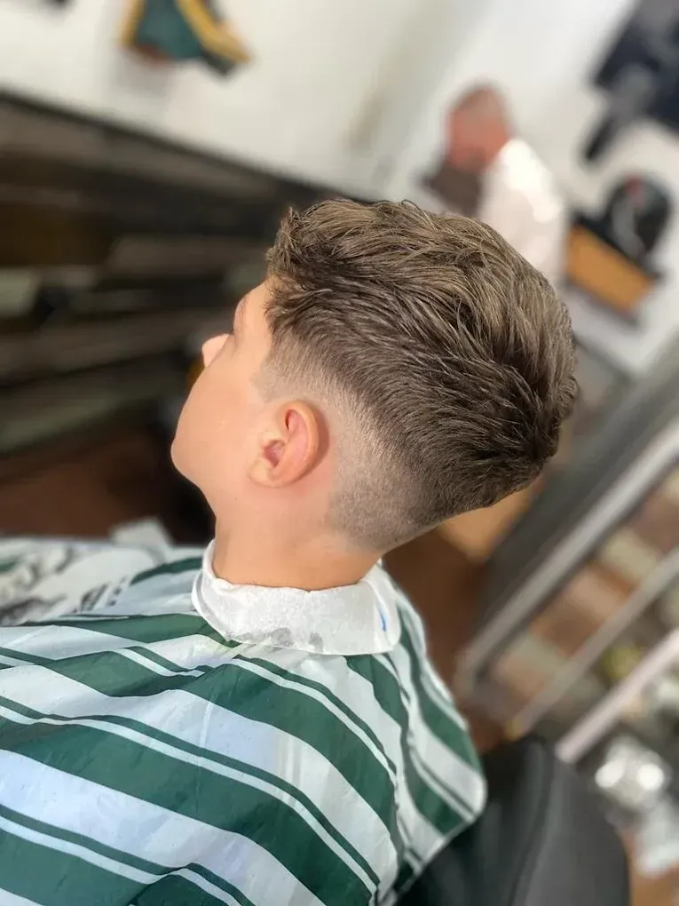 Niño con cabello texturizado y lados descoloridos, vistiendo una capa de barbero a rayas en un salón.