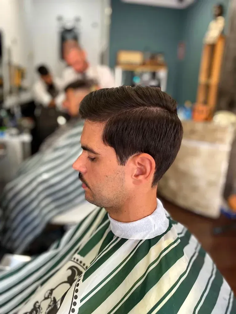 Hombre de pelo oscuro y bigote en una barbería, arreglándose el pelo. Lleva una capa a rayas.