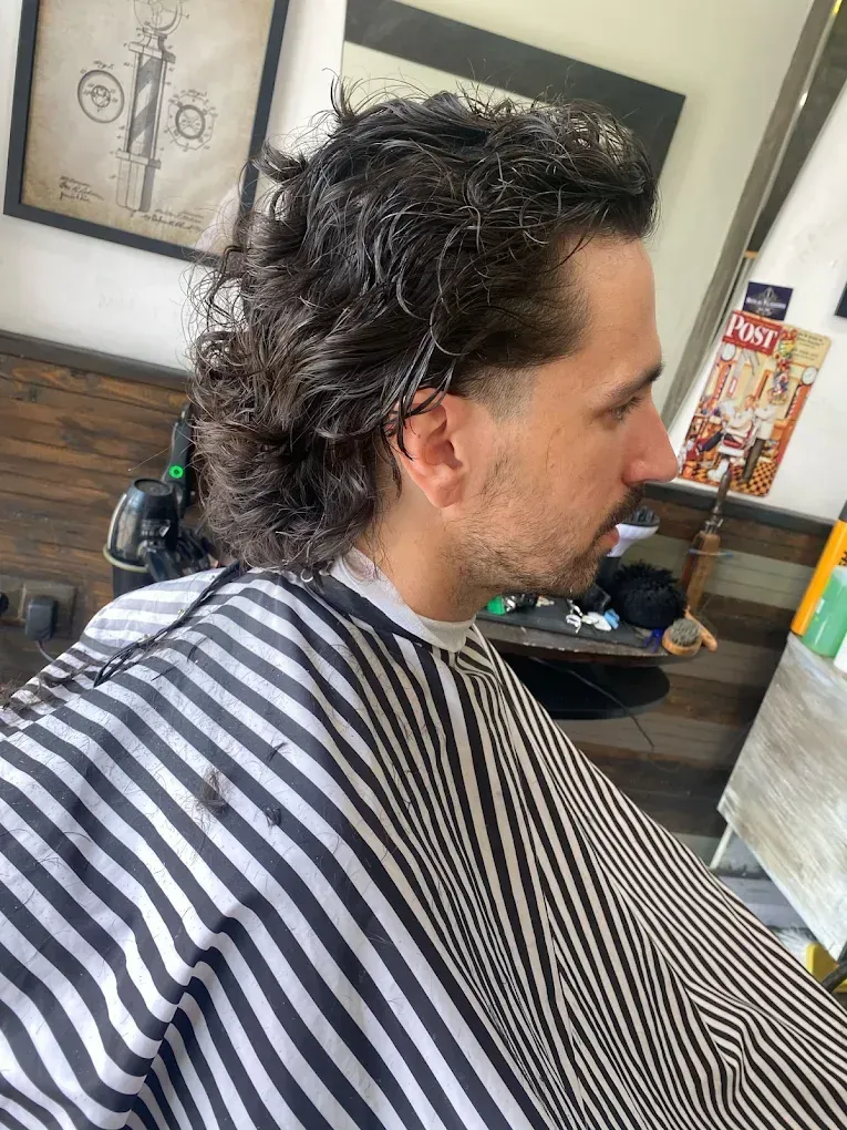 Hombre con cabello oscuro y ondulado con capa de barbero; vista lateral, entorno de barbería.