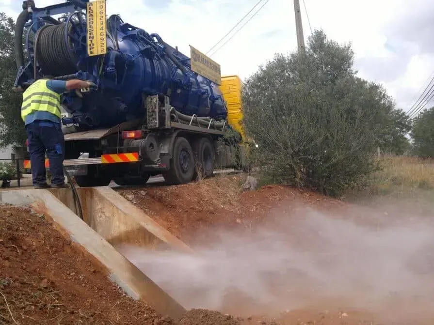 Caminhão-tanque despejando água barrenta em uma vala à beira da estrada enquanto um trabalhador está ao lado.
