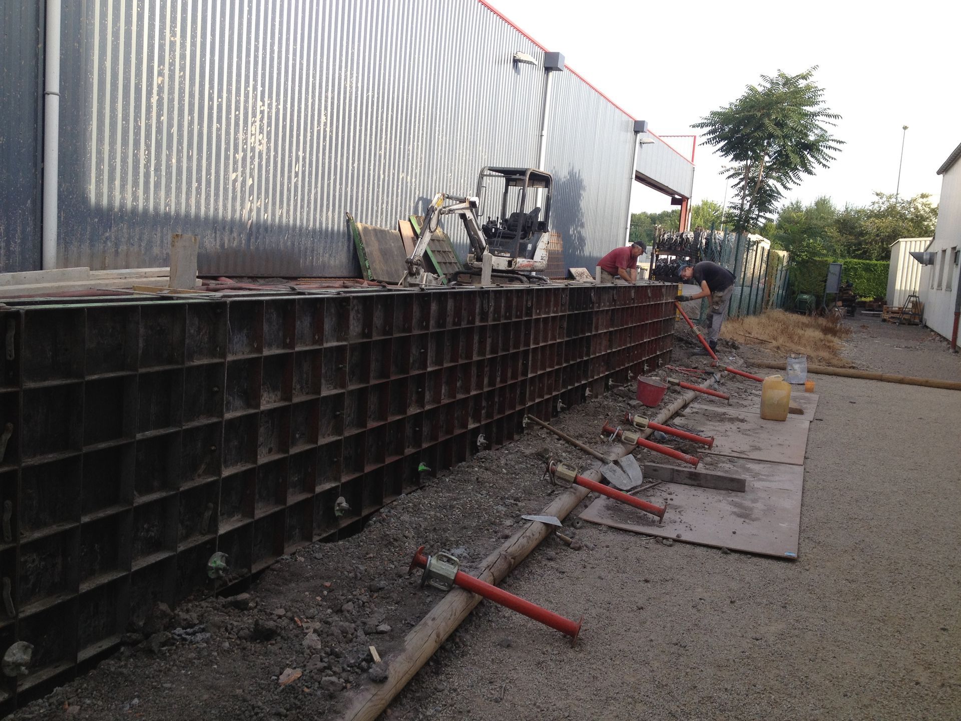 Chantier de construction avec coffrages métalliques pour un mur, sol gravillonné, bâtiment en tôle ondulée et ouvriers.