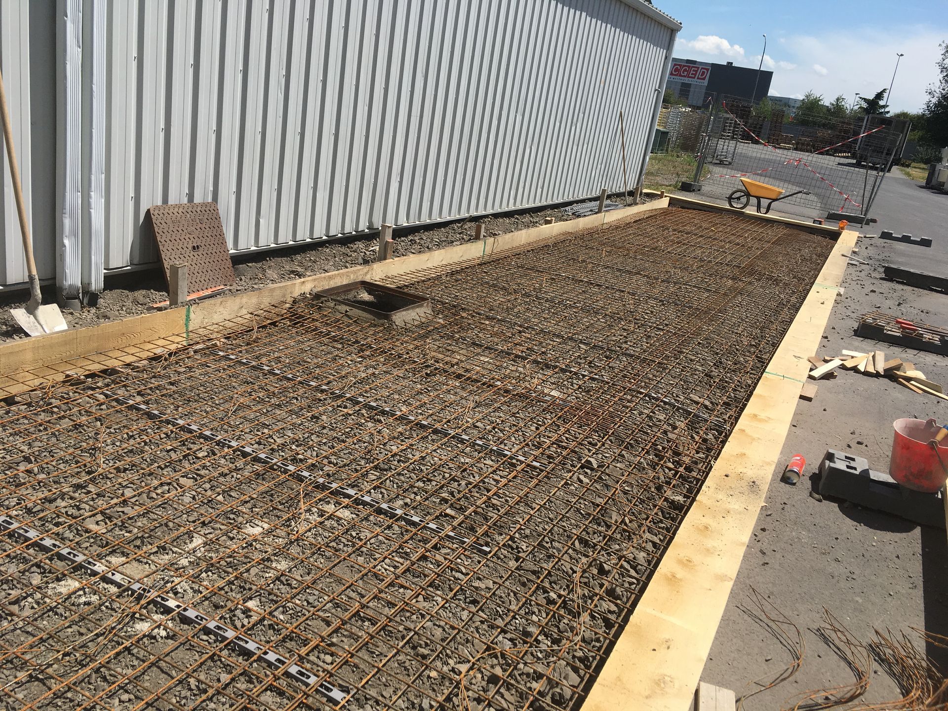 Dalle de béton en construction à côté d'un bâtiment, avec armatures, gravier et coffrages en bois.