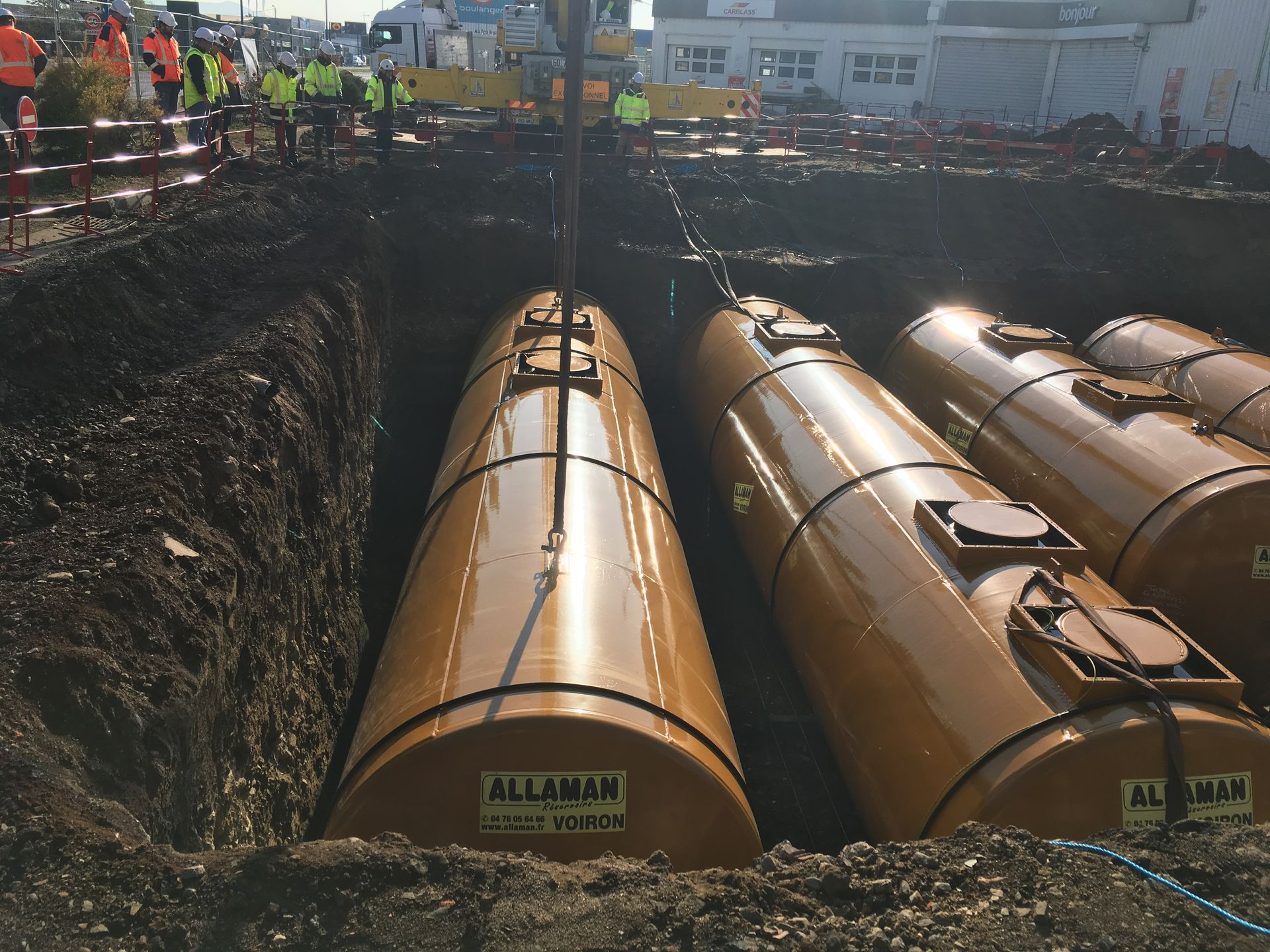 Une grande grue descend deux réservoirs de stockage souterrains cylindriques beiges dans une profonde tranchée
