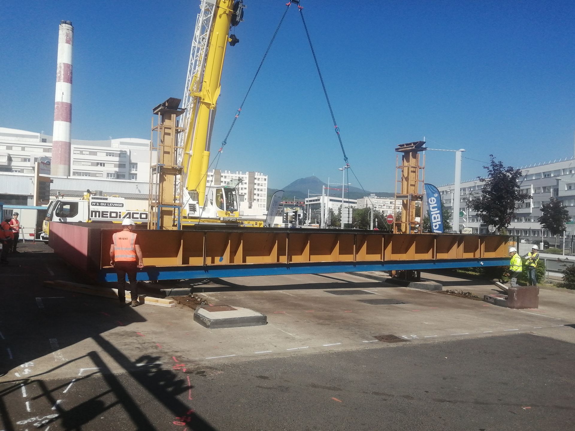 Sous un ciel bleu dégagé, une grue jaune soulève une longue poutre d'acier sur un chantier de construction industrielle.