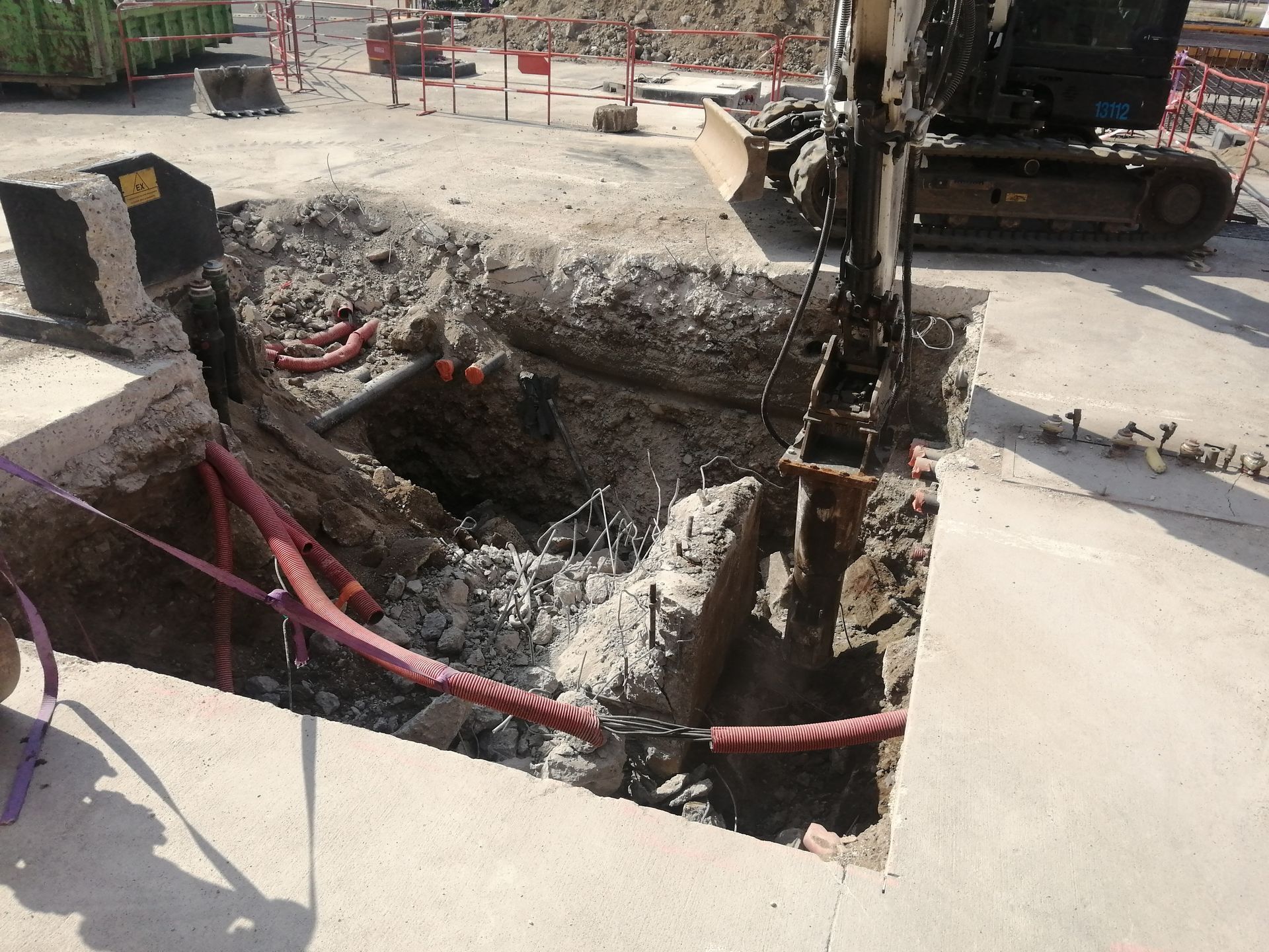 Chantier : Une excavatrice creuse dans une zone en béton, mettant à nu des câbles électriques.