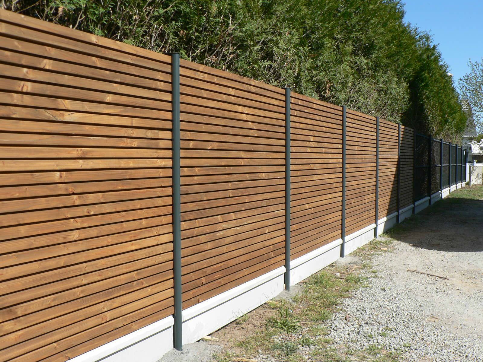 Clôture en lattes horizontales de bois avec poteaux foncés et base en béton clair, à côté de gravier.