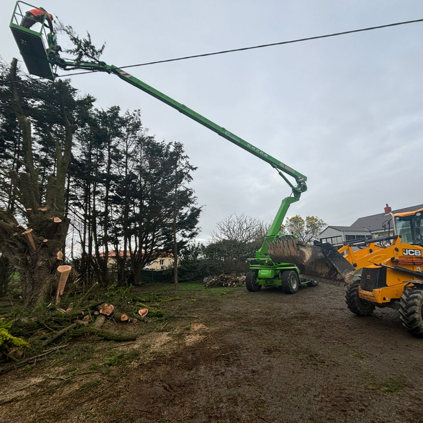 Un ouvrier, dans une nacelle verte, élague un arbre, tandis qu'une pelleteuse jaune et des branches coupées jonchent le sol.