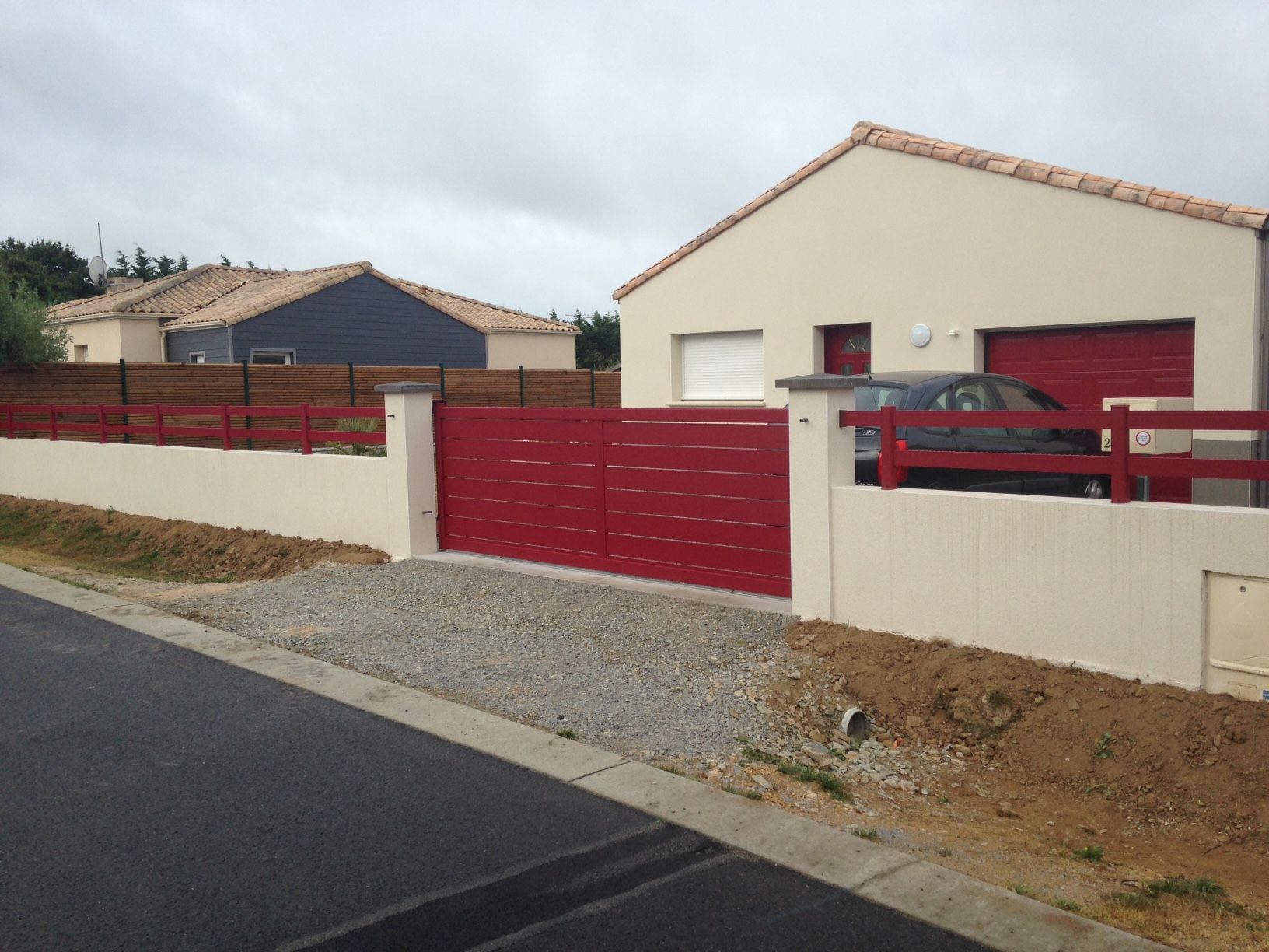 Portail et clôture rouges devant une maison beige avec une porte de garage rouge, dans une rue sous un ciel nuageux.