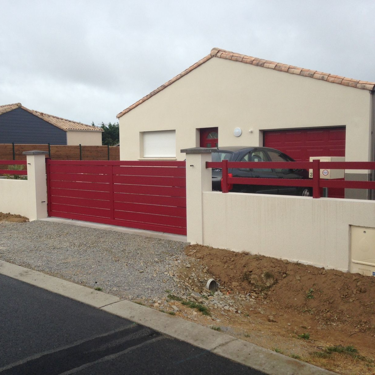 Portail et clôture rouges devant une maison beige avec garage ; allée de gravier gris et voiture noire à l'intérieur.