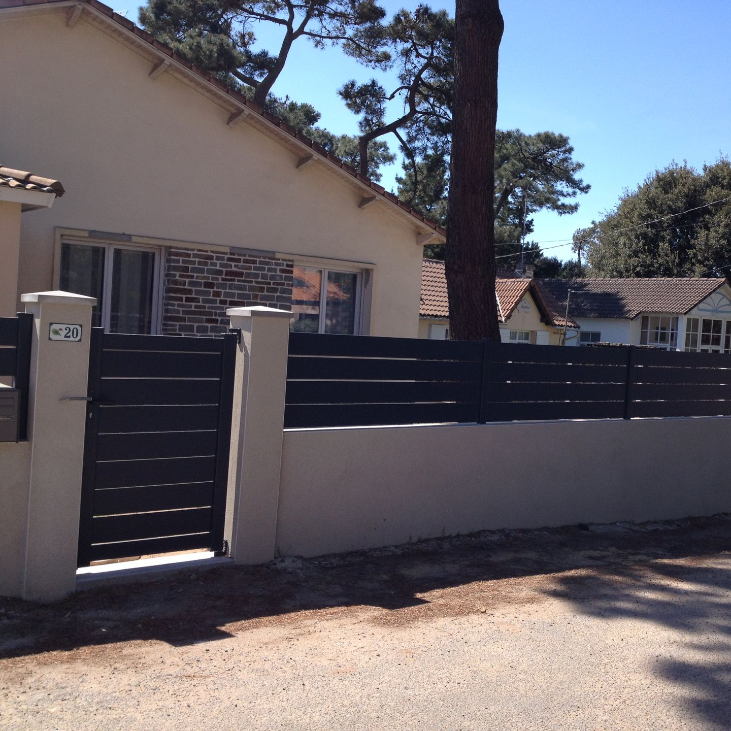 Maison beige avec clôture et portail noirs à l'avant ; journée ensoleillée.