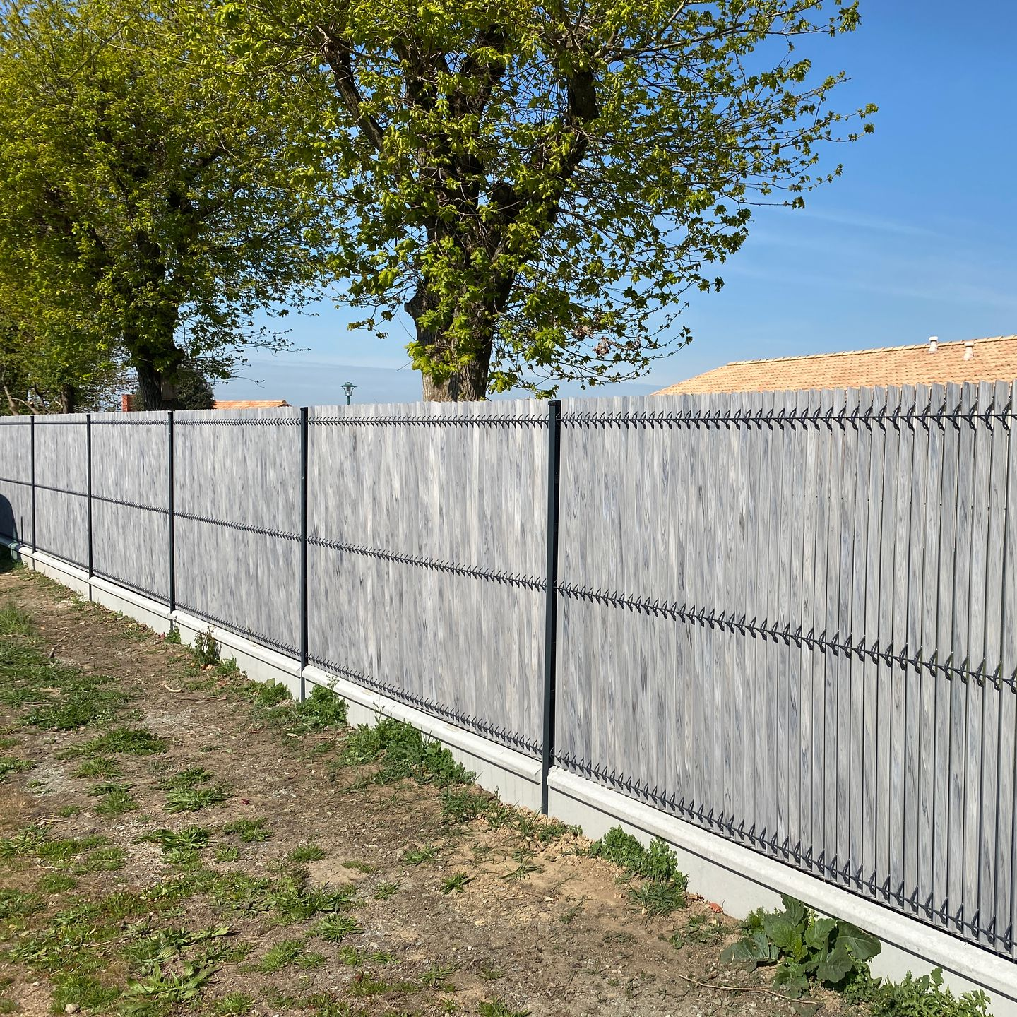 Clôture grise à claire-voie avec poteaux noirs, devant des arbres, par une journée ensoleillée.