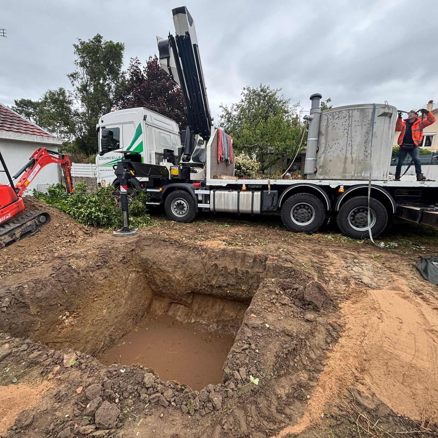 Chantier : camion avec grue, trou carré creusé, petite excavatrice, personne en gilet orange.