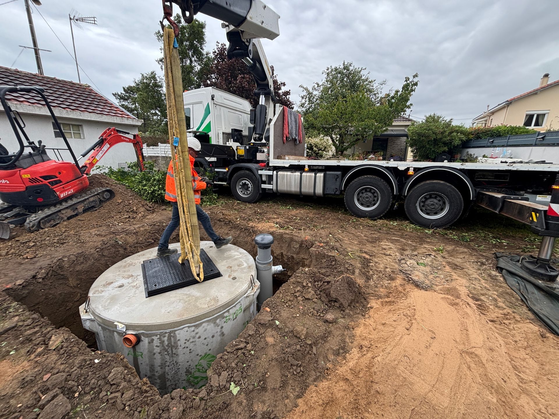 Une grue descend une fosse septique en béton dans un trou. Un ouvrier en gilet orange guide le chantier.