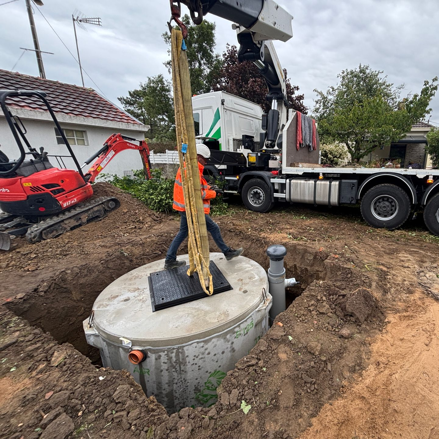 Une grue descend une fosse septique en béton dans un trou creusé. Un ouvrier en gilet orange guide la fosse. 