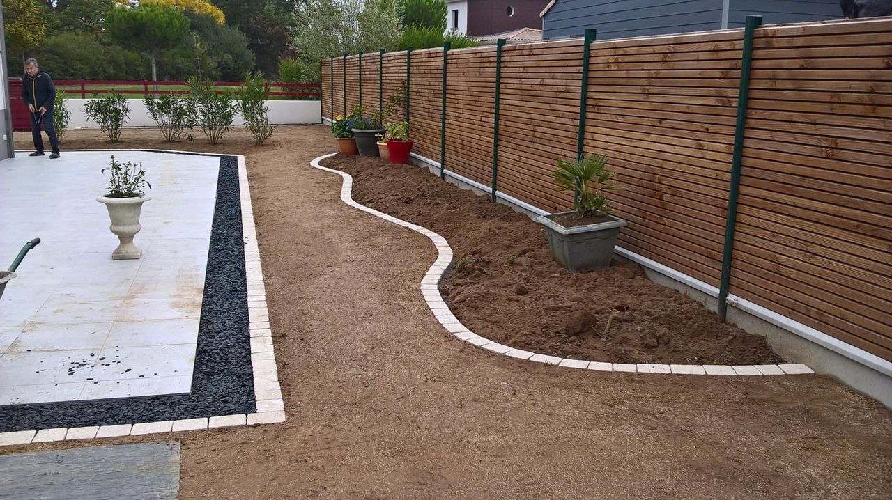 Une cour arrière avec une terrasse, un chemin de gravier, une clôture décorative et une personne qui marche.