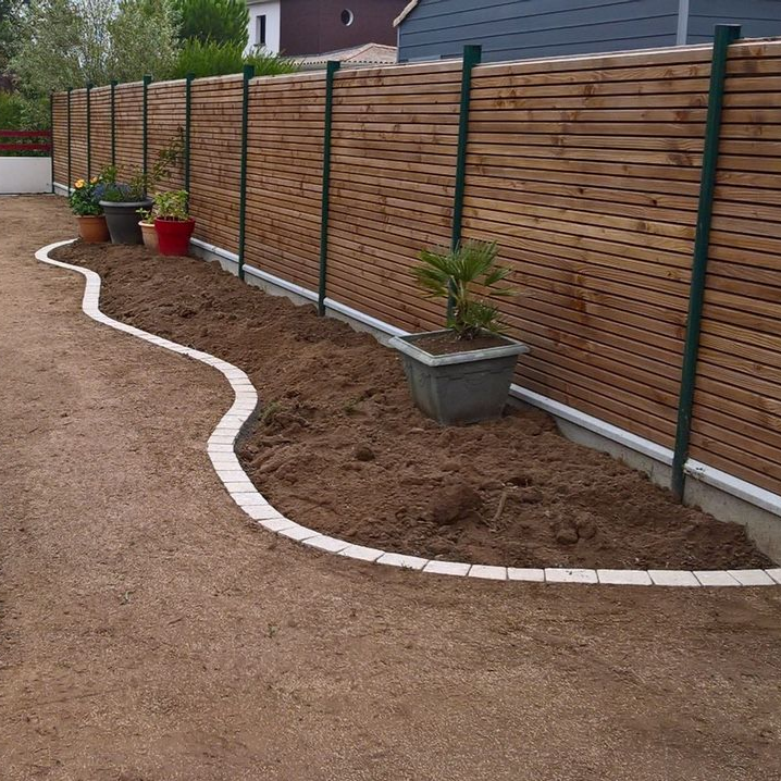 Clôture en bois brun avec des poteaux verts, bordée d'un rebord en pierre ondulée et de terre nue.