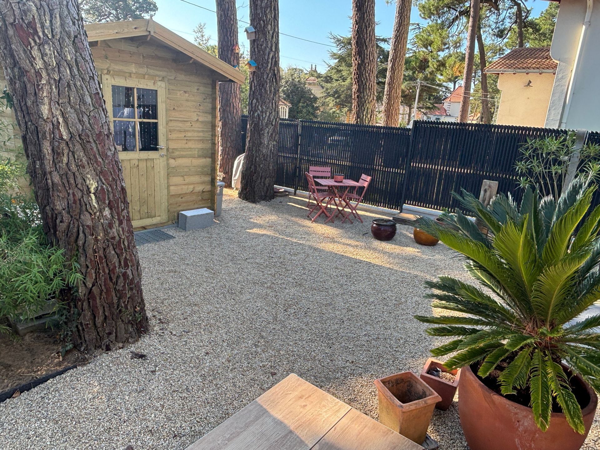 Une cour gravillonnée avec un petit abri de jardin en bois, des meubles de terrasse et des plantes en pot.
