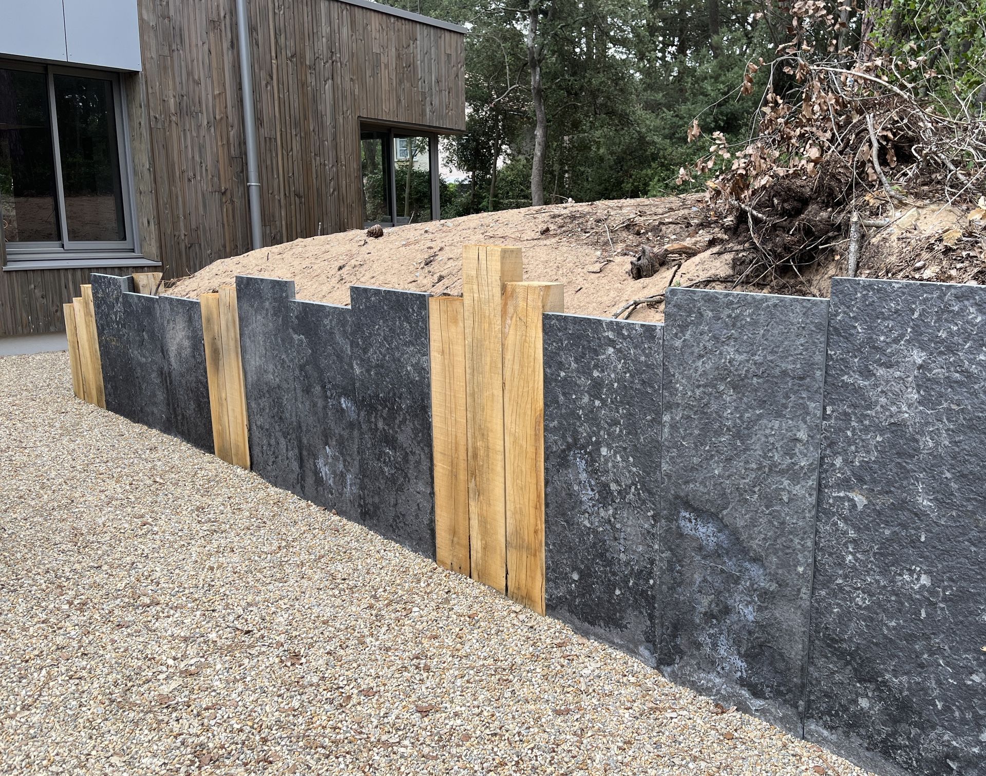 Mur de soutènement incurvé en blocs de pierre noire et supports en bois, gravier et sable dans un cadre boisé.