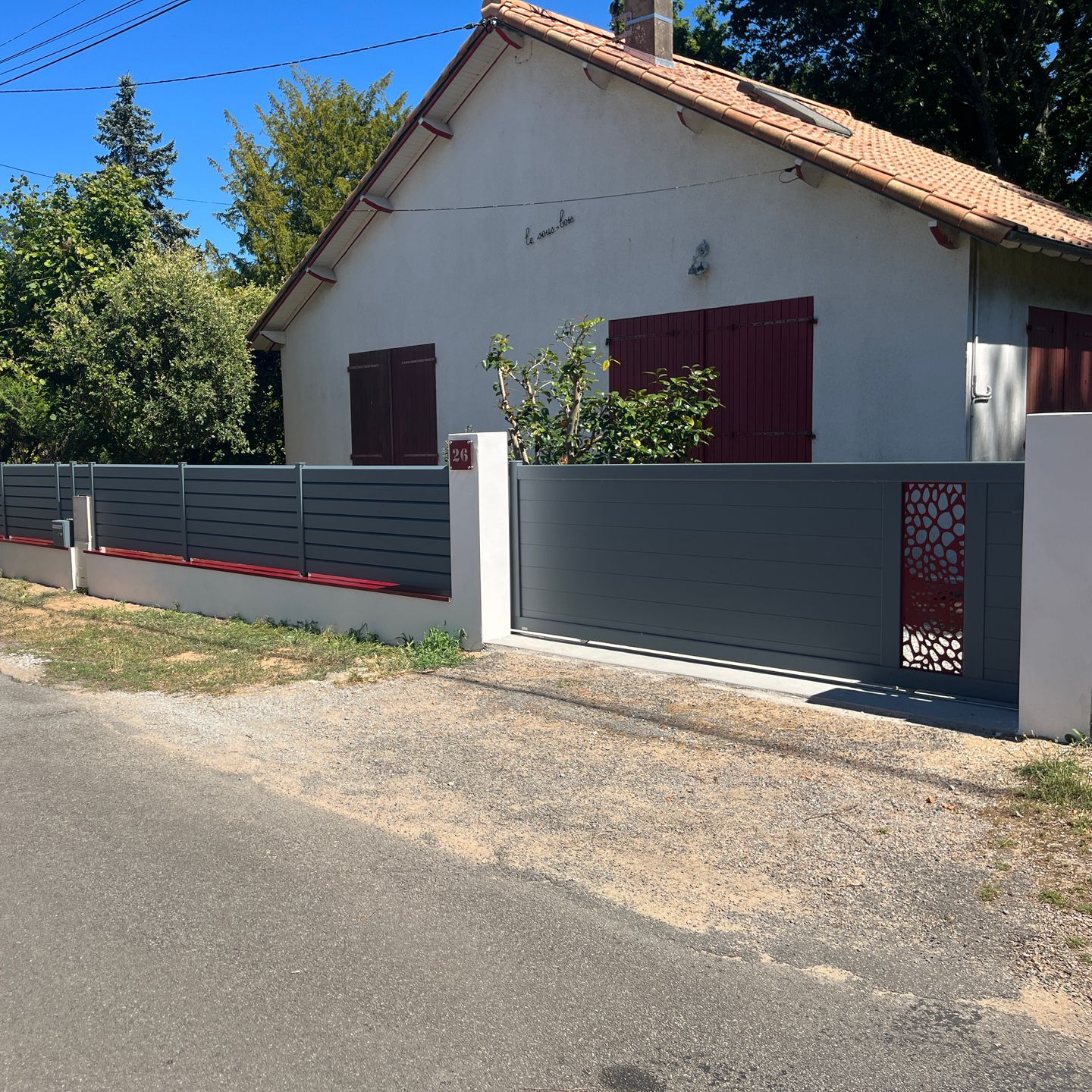 Clôture et portail gris devant une maison blanche avec des volets rouges et un panneau décoratif rouge sur le portail.