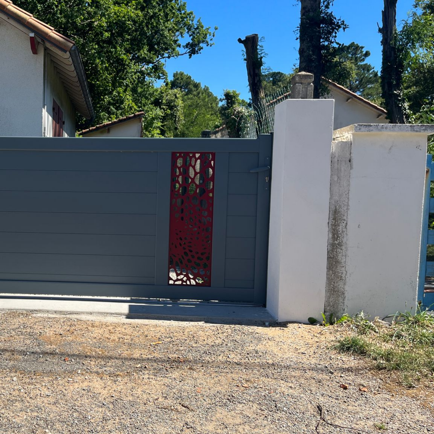 Portail d'allée gris avec panneau décoratif rouge, piliers blancs, gravier et arbres.