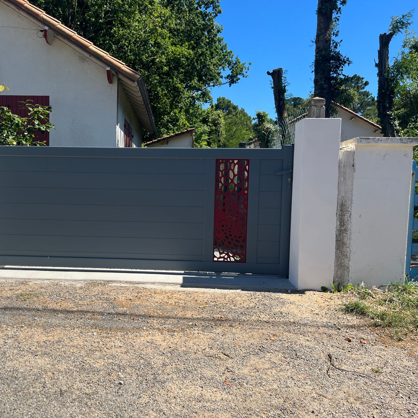 Portail coulissant gris avec panneau décoratif rouge et colonnes blanches devant une maison.