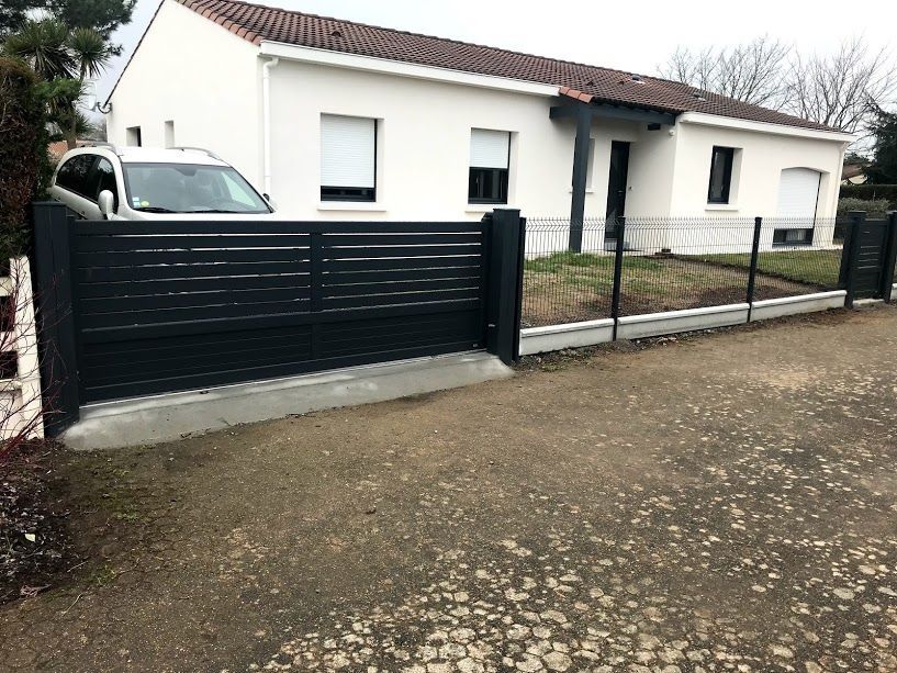 Portail métallique gris et rouge devant un pilier blanc et une maison, sol gravillonné, ciel bleu.