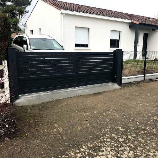 Portail en métal noir devant une maison blanche, une voiture étant partiellement visible.