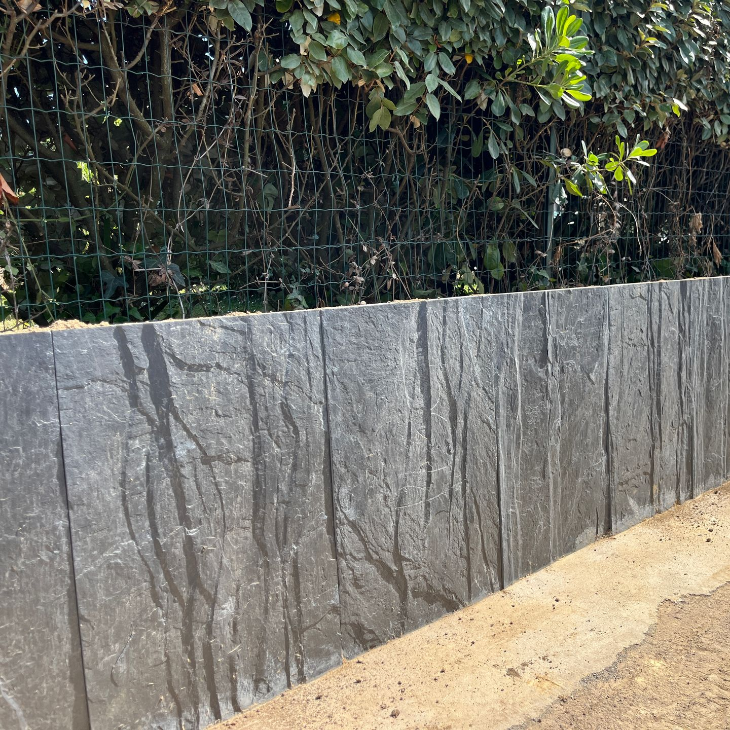 Mur de soutènement en pierre grise à surface texturée, adossé à une haie et une clôture vertes.