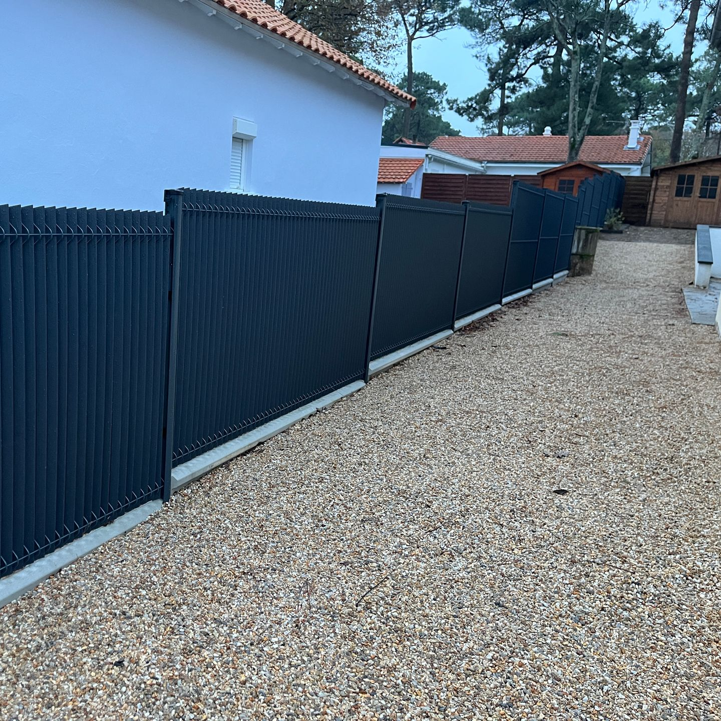 Clôture d'intimité bleu foncé le long d'un chemin de gravier, à côté d'une maison bleu pâle et d'autres habitations.