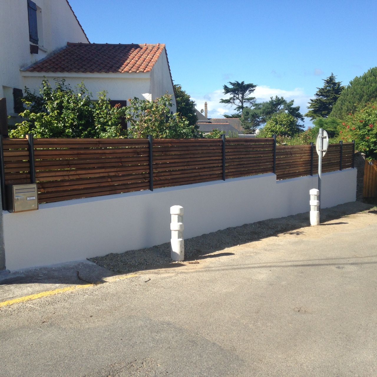 Un mur blanc surmonté d'une clôture en bois brun, au bord d'une route.