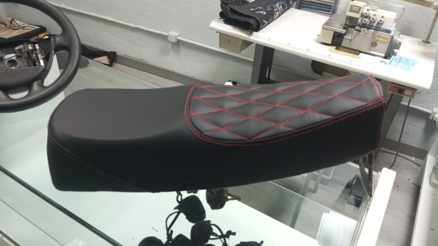 Asiento de motocicleta negro acolchado con costuras rojas expuesto sobre un mostrador de cristal.