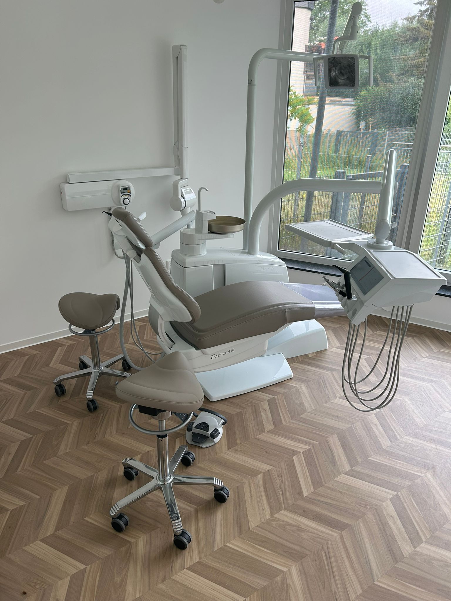 Zahnarztpraxis mit Stuhl, Instrumenten und zwei beigen Rollhockern. Fenster im Hintergrund.