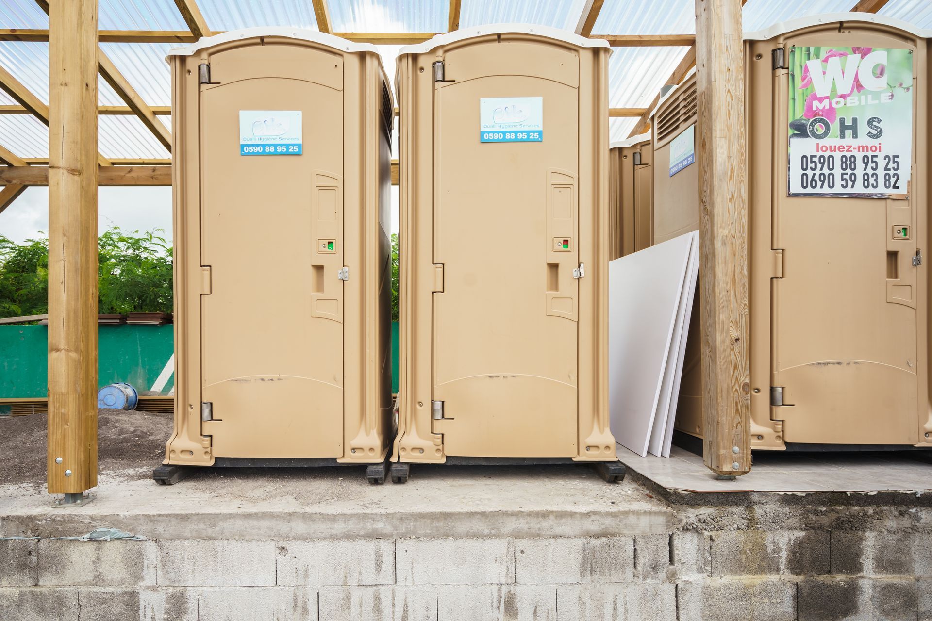 Deux toilettes portables beiges à côté d'une structure en bois sur une dalle de béton à l'extérieur